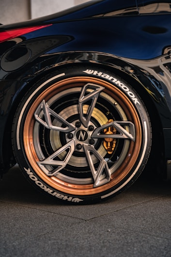 A close-up of a car's wheel featuring a high-performance tire with the brand name 'Hankook' visible. The wheel has a sleek, modern design with a metallic finish and copper-colored rim. The brake caliper is visible inside, adding to the sporty aesthetic. The car's body is dark and glossy, reflecting the surroundings.