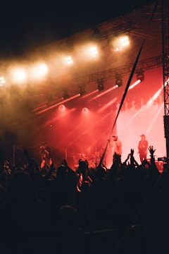 A dynamic concert scene with performers on stage under bright lights. The crowd is actively engaged, with hands raised, while the stage features musicians and vocalists. Warm lighting enhances the energetic atmosphere.