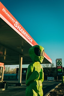 A large green alien statue stands next to a gas station with a red and white sign reading 'Diamond Shamrock.' The bright blue sky contrasts with the vivid colors of the statue and the surrounding environment. A fuel price sign is visible in the background, along with some trees and buildings.