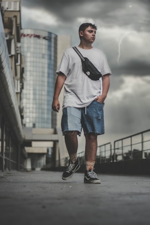A young man stands on a concrete pathway in an urban environment. He is wearing a white oversized t-shirt, blue denim shorts, and black lace-up shoes. He has a black crossbody bag slung over his shoulder. In the background, there is a tall glass building and overcast, stormy skies with a visible lightning bolt.