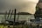A trailer is parked by a chain-link fence beside a turbulent body of water. The scene includes a hill in the background and some boats near a dock. The sky appears overcast and hazy, contributing to a stormy atmosphere.