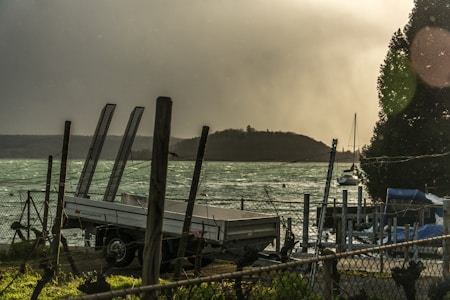 A trailer is parked by a chain-link fence beside a turbulent body of water. The scene includes a hill in the background and some boats near a dock. The sky appears overcast and hazy, contributing to a stormy atmosphere.