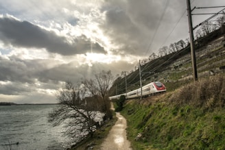 A high-speed train travels along a railway track bordering a river. The sky is overcast with numerous clouds, allowing rays of sunlight to peak through. The railway is flanked by a steep hillside covered with sparse vegetation.