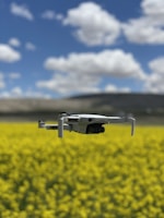 A drone hovers in the foreground over a vibrant yellow field, likely composed of flowering plants. In the background, a blue sky with scattered white clouds stretches above distant hills or mountains.