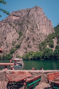 A scenic view of a tranquil river surrounded by steep, rocky mountains with patches of greenery. A tour boat is docked on the river with people visible on the dock and nearby. In the foreground, wooden picnic tables and benches are set on a stone terrace, offering a relaxed spot to enjoy the surroundings.