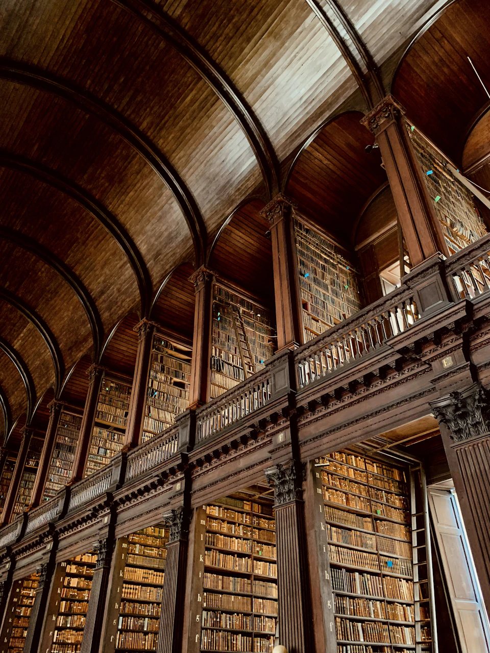 Old library with high ceiling and wooden beams features numerous stacked bookshelves upstairs