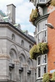An architectural scene featuring a classic stone building with ornate arches and decorative balustrades. Adjacent is a brick building with bay windows and plants draping from window ledges. A satellite dish and a streetlamp are visible against a backdrop of a partly cloudy sky.