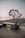 A monument in the shape of the African continent stands on a rocky coastline at sunset. The structure features interconnected lines and sits on a dark base inscribed with text, which includes coordinates and the name Cap Angela.