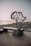 A monument in the shape of the African continent stands on a rocky coastline at sunset. The structure features interconnected lines and sits on a dark base inscribed with text, which includes coordinates and the name Cap Angela.