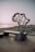 A monument in the shape of the African continent stands on a rocky coastline at sunset. The structure features interconnected lines and sits on a dark base inscribed with text, which includes coordinates and the name Cap Angela.