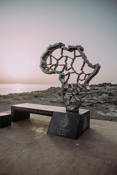 A monument in the shape of the African continent stands on a rocky coastline at sunset. The structure features interconnected lines and sits on a dark base inscribed with text, which includes coordinates and the name Cap Angela.