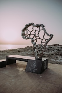 A monument in the shape of the African continent stands on a rocky coastline at sunset. The structure features interconnected lines and sits on a dark base inscribed with text, which includes coordinates and the name Cap Angela.