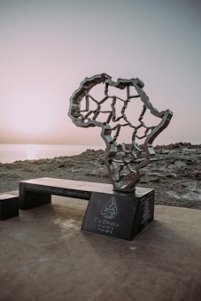 A monument in the shape of the African continent stands on a rocky coastline at sunset. The structure features interconnected lines and sits on a dark base inscribed with text, which includes coordinates and the name Cap Angela.
