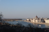 A panoramic view of a river with a grand historic building, likely a parliament, located along its banks. A bridge spans the river, with a couple of boats visible on the water. The buildings appear majestic, with intricate architectural details, and the scene is framed by trees in the foreground.