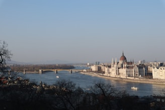 A panoramic view of a river with a grand historic building, likely a parliament, located along its banks. A bridge spans the river, with a couple of boats visible on the water. The buildings appear majestic, with intricate architectural details, and the scene is framed by trees in the foreground.