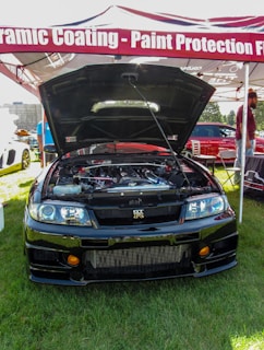 A black sports car with its hood open is displayed under a canopy that advertises ceramic coating and paint protection. The car's engine is visible, indicating a showcase or auto event. Surrounding the car are people and other vehicles, with a grassy area underfoot.