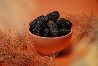 A bowl filled with dark, wrinkled dates is placed on an orange surface, surrounded by dried plants with a soft focus.