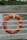 A bright orange lifebuoy is hanging on a railing with a body of water and a green landscape visible in the background. The lifebuoy is secured with ropes and has white reflective strips.