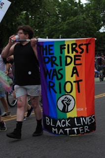 A person walks down a street holding a colorful banner with rainbow colors prominently displayed. The banner reads 'The First Pride Was a Riot,' along with 'Black Lives Matter' and features a black fist symbol. The person appears to be participating in a parade or protest, as partially visible people and signs can be seen in the background.