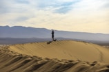 A person is standing on top of a large sand dune, holding a camera to capture the vast landscape. The horizon features a range of distant mountains under a partly cloudy sky.