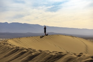 A person is standing on top of a large sand dune, holding a camera to capture the vast landscape. The horizon features a range of distant mountains under a partly cloudy sky.