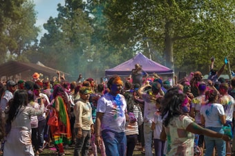 A large group of people gathered outdoors during a colorful festival, with vibrant powders covering their clothes, faces, and surroundings. Lush green trees are in the background, and there is a purple tent in the center. The crowd is lively, with many people raising their hands and others taking photos.