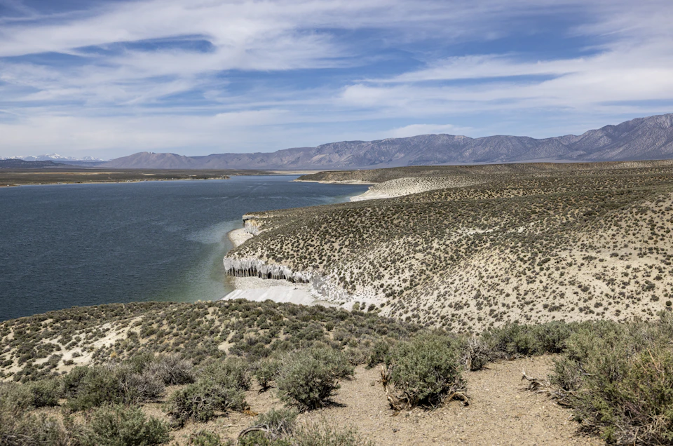 The image depicts a serene landscape with a large body of water, possibly a lake or a river, nestled between two mountains on the left side and surrounded by lush green vegetation on the right side. The sky above is a clear blue, dotted with fluffy white clouds that add to the overall tranquility of the scene.

The perspective of the image is from a high vantage point, looking down upon the landscape. This viewpoint provides a comprehensive view of the natural beauty and grandeur of the area.