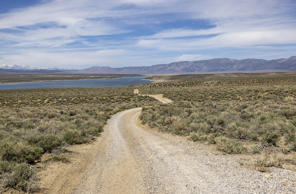 The image depicts a winding dirt road cutting through a vast, open landscape. The road is surrounded by tall grass and shrubs on both sides, with mountains rising in the distance beyond it. The sky above is clear blue with scattered clouds.