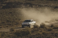 A white flatbed truck loaded with equipment, driving along a dusty road.