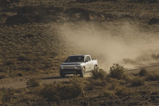 A white flatbed truck loaded with equipment, driving along a dusty road.