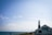 A lighthouse stands near a restaurant called Lobster Pot along the coastline. The sky is clear with a few clouds, and the sea is visible to the left. Several people are gathered near the restaurant and the shore. Seagulls are flying above, adding to the coastal scene.