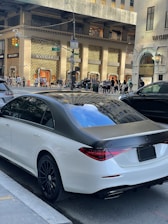 A luxury sedan is parked on a city street in front of high-end retail stores like BVLGARI. Pedestrians walk along the sidewalk and cars are visible on the street. The environment suggests a busy urban area with upscale shopping and modern architecture.