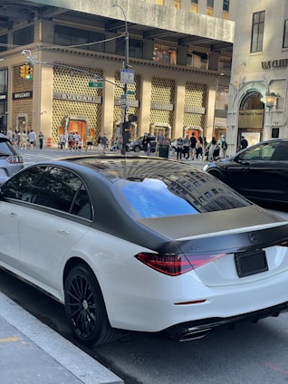 A luxury sedan is parked on a city street in front of high-end retail stores like BVLGARI. Pedestrians walk along the sidewalk and cars are visible on the street. The environment suggests a busy urban area with upscale shopping and modern architecture.