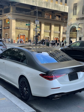 A luxury sedan is parked on a city street in front of high-end retail stores like BVLGARI. Pedestrians walk along the sidewalk and cars are visible on the street. The environment suggests a busy urban area with upscale shopping and modern architecture.