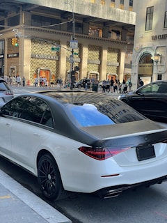 A luxury sedan is parked on a city street in front of high-end retail stores like BVLGARI. Pedestrians walk along the sidewalk and cars are visible on the street. The environment suggests a busy urban area with upscale shopping and modern architecture.