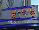 A close-up of a sign with red neon letters spelling 'RADIO CITY' and smaller gold letters below spelling 'MUSIC HALL'. The sign features horizontal blue lighting stripes and is set against a grey building façade.