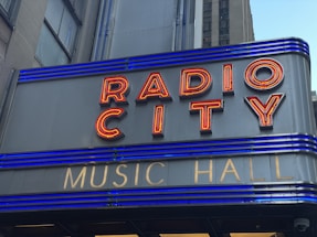 A close-up of a sign with red neon letters spelling 'RADIO CITY' and smaller gold letters below spelling 'MUSIC HALL'. The sign features horizontal blue lighting stripes and is set against a grey building façade.