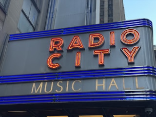 A close-up of a sign with red neon letters spelling 'RADIO CITY' and smaller gold letters below spelling 'MUSIC HALL'. The sign features horizontal blue lighting stripes and is set against a grey building façade.