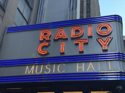 A close-up of a sign with red neon letters spelling 'RADIO CITY' and smaller gold letters below spelling 'MUSIC HALL'. The sign features horizontal blue lighting stripes and is set against a grey building façade.