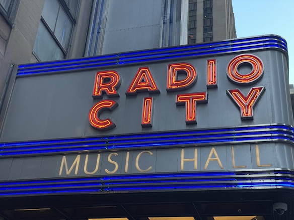 A close-up of a sign with red neon letters spelling 'RADIO CITY' and smaller gold letters below spelling 'MUSIC HALL'. The sign features horizontal blue lighting stripes and is set against a grey building façade.