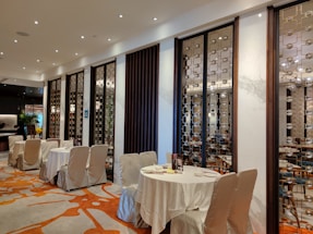 A stylish and upscale restaurant interior with neatly arranged tables covered in white cloth. The walls are adorned with decorative panels featuring intricate geometric patterns. Warm lighting from numerous small ceiling lights creates an inviting ambiance, and the wooden furniture adds an element of elegance. Subtle green plants are visible in the background.