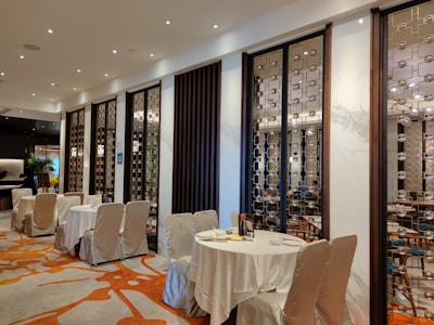 A stylish and upscale restaurant interior with neatly arranged tables covered in white cloth. The walls are adorned with decorative panels featuring intricate geometric patterns. Warm lighting from numerous small ceiling lights creates an inviting ambiance, and the wooden furniture adds an element of elegance. Subtle green plants are visible in the background.
