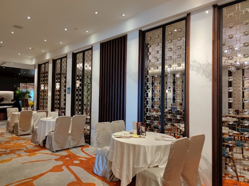 A stylish and upscale restaurant interior with neatly arranged tables covered in white cloth. The walls are adorned with decorative panels featuring intricate geometric patterns. Warm lighting from numerous small ceiling lights creates an inviting ambiance, and the wooden furniture adds an element of elegance. Subtle green plants are visible in the background.