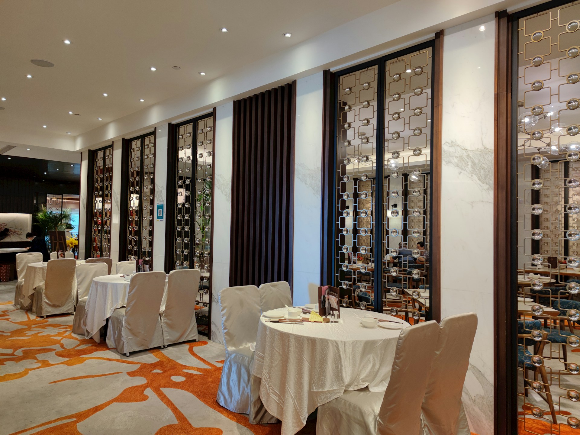 A stylish and upscale restaurant interior with neatly arranged tables covered in white cloth. The walls are adorned with decorative panels featuring intricate geometric patterns. Warm lighting from numerous small ceiling lights creates an inviting ambiance, and the wooden furniture adds an element of elegance. Subtle green plants are visible in the background.