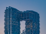 A modern high-rise building with a unique architectural design featuring two towers connected by a bridge at the upper levels. The structure displays a series of staggered balconies, giving it a layered appearance. The overall style is contemporary, with a stark, geometric facade.