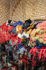 A large pile of colorful yarns and threads is stacked against a brick wall with an arched pattern. The yarns are tangled and come in a variety of colors including red, blue, yellow, and orange, creating a vibrant and somewhat chaotic scene.