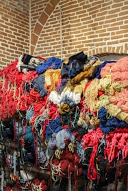 A large pile of colorful yarns and threads is stacked against a brick wall with an arched pattern. The yarns are tangled and come in a variety of colors including red, blue, yellow, and orange, creating a vibrant and somewhat chaotic scene.