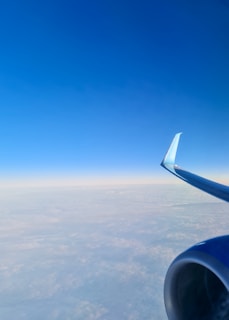 An airplane wing and engine are visible against a vast sky, with a clear blue gradient extending upward. Below, a layer of clouds adds texture to the expansive view, offering a sense of altitude and open space.