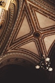Intricate ceiling design featuring ornate patterns and detailed artwork. The design includes symmetrical geometric shapes adorned with floral motifs and rich textures, accompanied by an elegant chandelier with multiple lights.