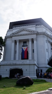 A grand, white neoclassical building with tall columns and a triangular pediment. A large flag is draped between two of the columns. People can be seen gathered at the entrance, which is framed by an archway. The structure is surrounded by a well-maintained lawn with trees and a notable rock in the foreground.